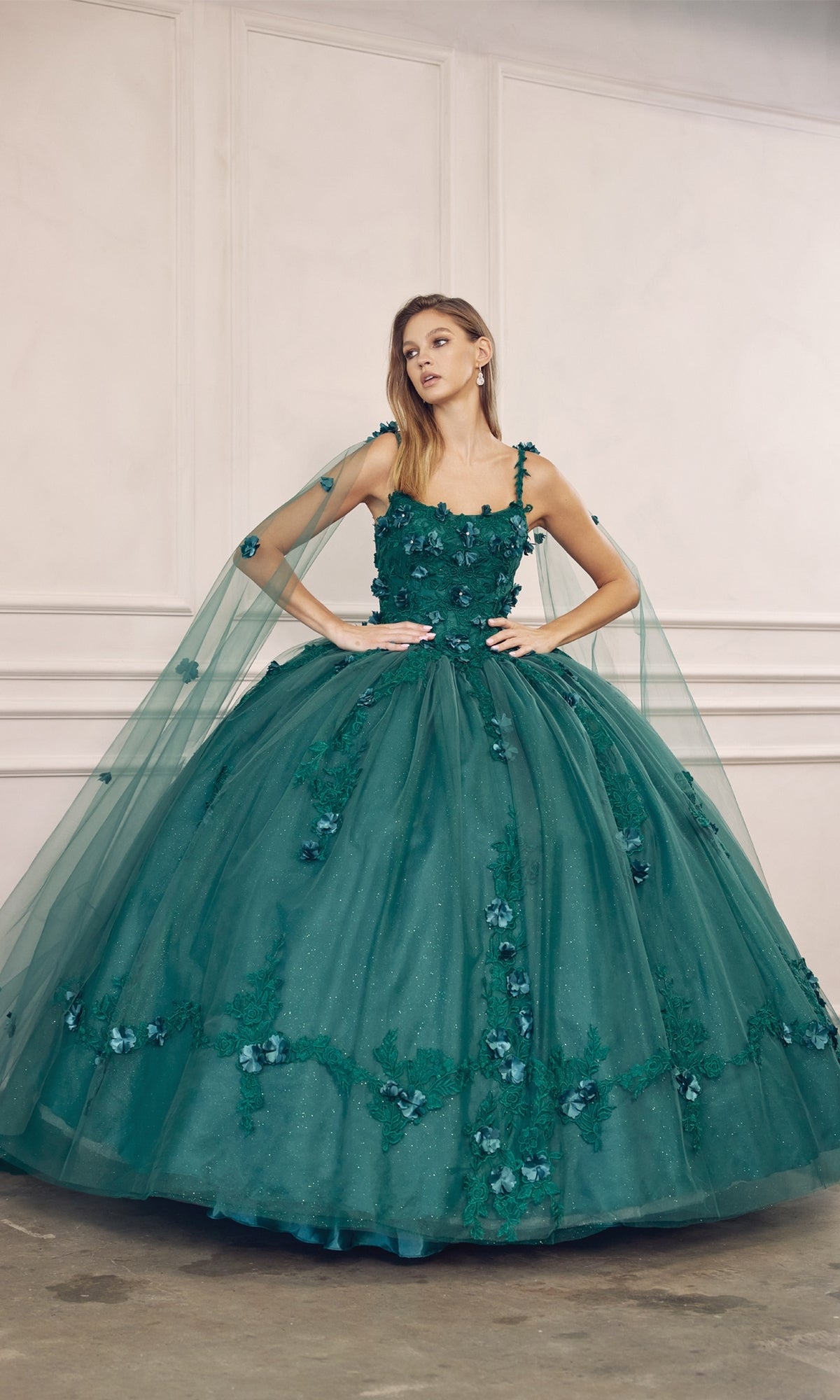 A woman wears the Juliet 1436 Glitter Quinceañera Dress with cape, featuring beaded embroidery, floral appliqués, a voluminous tulle skirt, and a matching sheer cape, set against a white-paneled wall.